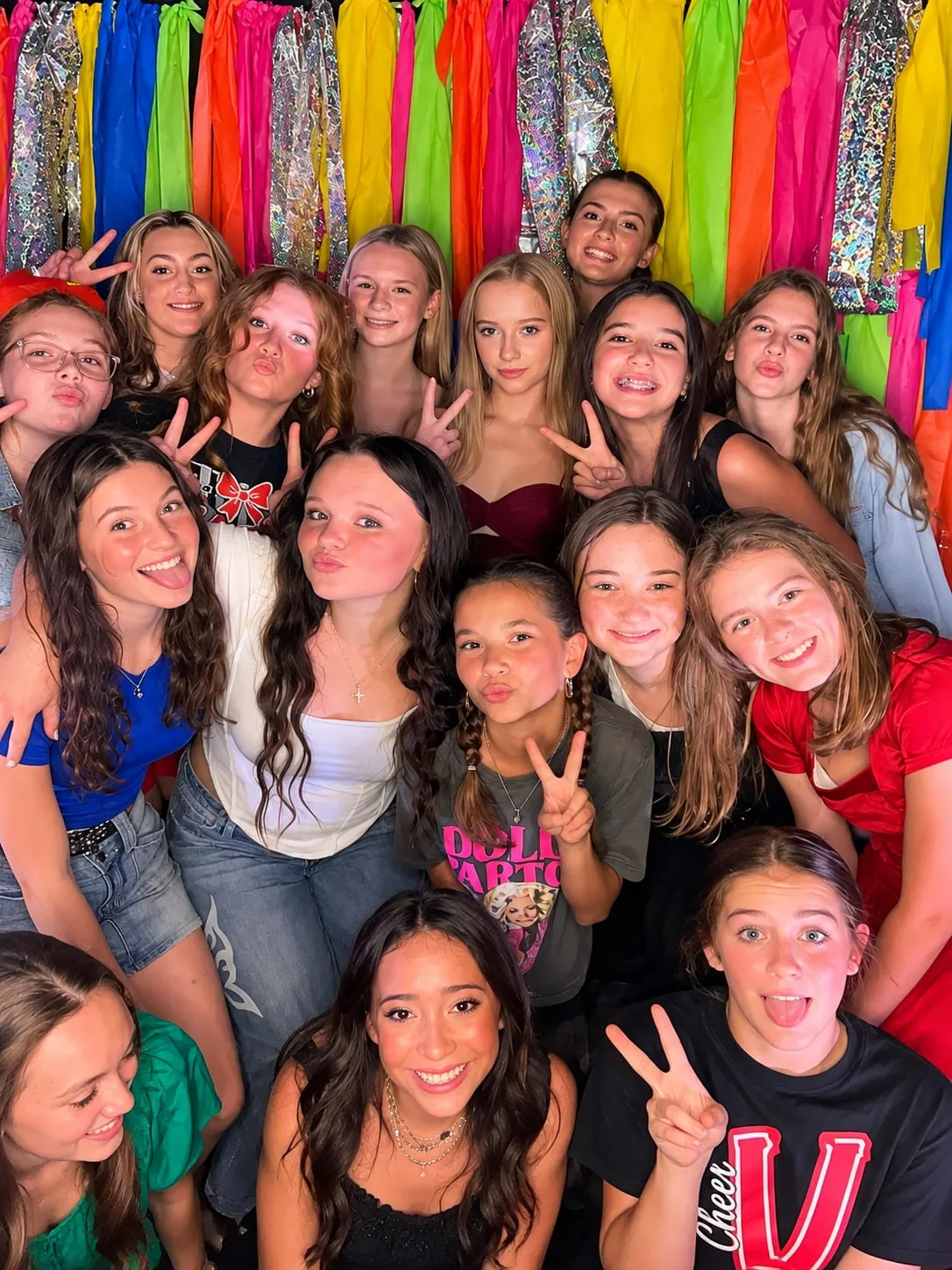 A large group of students packed into frame together in a school gym, pulling faces in front of a rainbow fringe wall