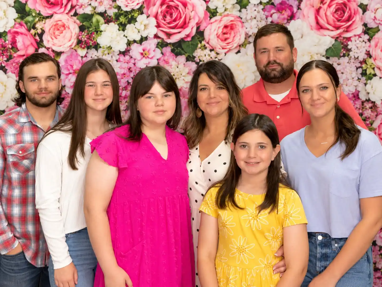 A family of six standing together in front of a wall of pink and cream roses