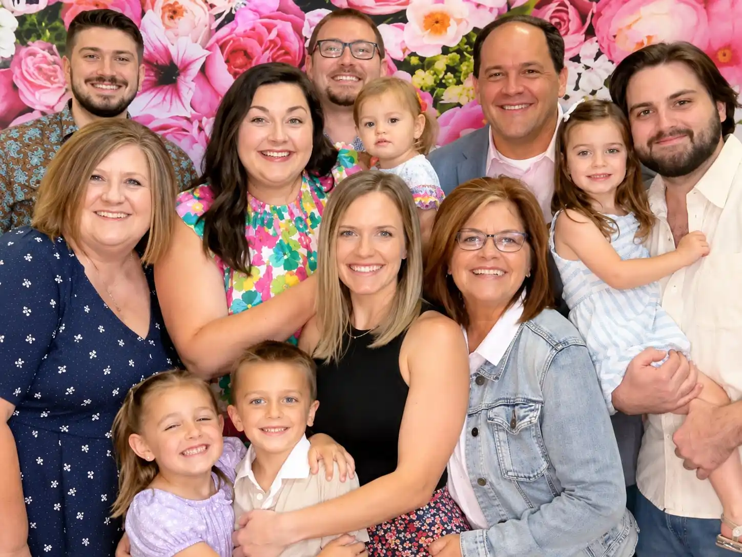 Three generations of a family crowded into one shot in front of a wall of pink roses, the youngest on a grandparent's hip