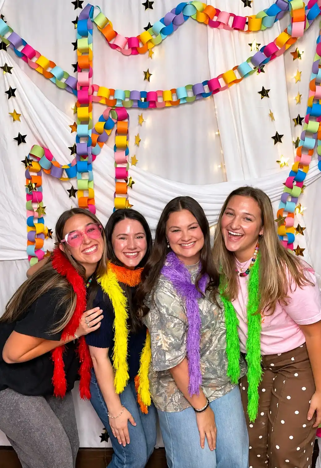 Four friends in feather boas laughing together in front of a white curtain strung with a paper chain garland