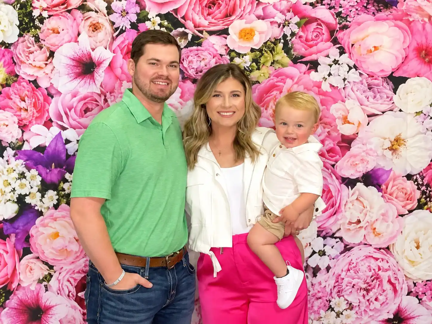 A couple holding their toddler, all three mid-laugh, against a wall of pink and white roses