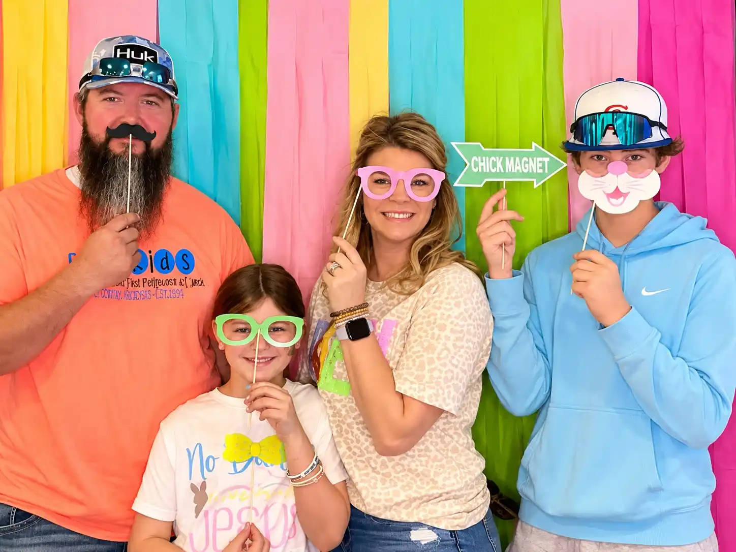 A family of four holding props and novelty glasses in front of a wall of bright pastel streamers