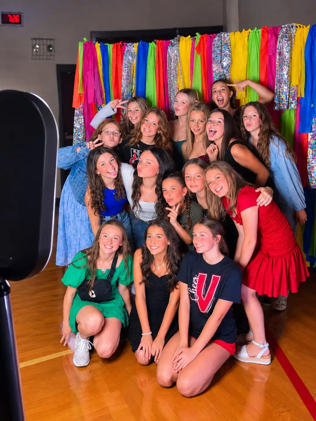 A cheer squad crowded into frame in a school gym, in front of a rainbow fringe wall hung with silver streamers