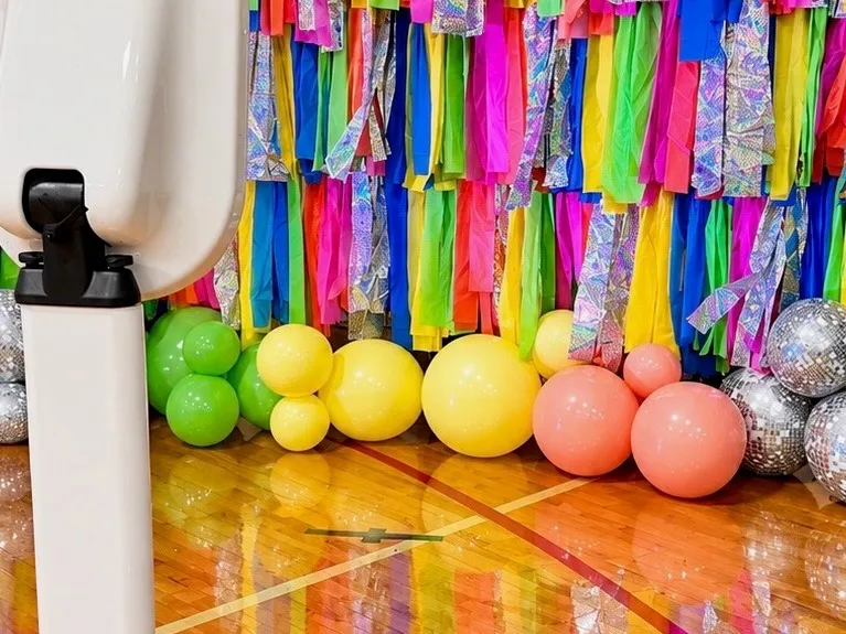 The selfie booth standing in position before an event, beside a balloon garland on a polished gym floor